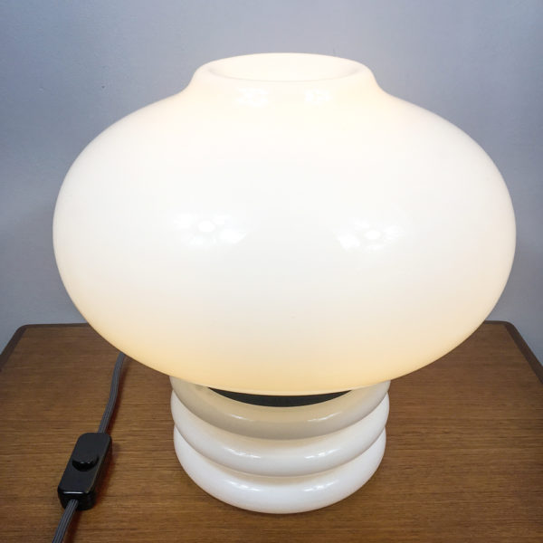 Für ein modernes zu Haues. Wer auf die Space Age Ära steht, sollte sich diese Lampe unbedingt für zu Hause kaufen. Die Tischlampe besteht komplett aus weissem, opakem Glas. Der obere Teil ist in der Form einer Blase gestaltet und wird durch einen schwarzen Metallring vom unteren Teil unterbrochen. Hier wird auch die Lampe geöffnet, wenn man das Leuchtmittel austauschen möchte. Der schwarze Metallring ist das Verbindungsstück zwischen dem Oberteil und Unterteil der Tischlampe. Der untere Teil der Tischlampe besteht ebenfalls aus weissem, opakem Glas und ist aus einer zylindrischen Form, die drei ringförmige Auswölbungen nach aussen hat. Die Gesamthöhe der Lampe beträgt 36cm. Die Kugel (Bubble) hat einen Durchmesser von 40cm. Der zylindrische Fuss mit den Auswölbungen hat einen Durchmesser von 20cm und eine Höhe von 16cm inkl. des schwarzen Metallringes. Dementsprechend ergibt sich eine Höhe von 20cm für die Blase, die den Lampenschirm darstellt.