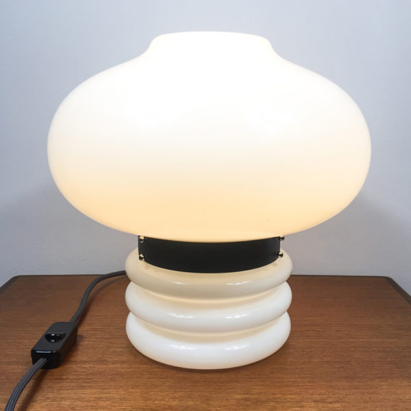 Für ein modernes zu Haues. Wer auf die Space Age Ära steht, sollte sich diese Lampe unbedingt für zu Hause kaufen. Die Tischlampe besteht komplett aus weissem, opakem Glas. Der obere Teil ist in der Form einer Blase gestaltet und wird durch einen schwarzen Metallring vom unteren Teil unterbrochen. Hier wird auch die Lampe geöffnet, wenn man das Leuchtmittel austauschen möchte. Der schwarze Metallring ist das Verbindungsstück zwischen dem Oberteil und Unterteil der Tischlampe. Der untere Teil der Tischlampe besteht ebenfalls aus weissem, opakem Glas und ist aus einer zylindrischen Form, die drei ringförmige Auswölbungen nach aussen hat. Die Gesamthöhe der Lampe beträgt 36cm. Die Kugel (Bubble) hat einen Durchmesser von 40cm. Der zylindrische Fuss mit den Auswölbungen hat einen Durchmesser von 20cm und eine Höhe von 16cm inkl. des schwarzen Metallringes. Dementsprechend ergibt sich eine Höhe von 20cm für die Blase, die den Lampenschirm darstellt.