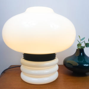 Für ein modernes zu Hause. Wer auf die Space Age Ära steht, sollte sich diese Lampe unbedingt für zu Hause kaufen. Die Tischlampe besteht komplett aus weissem, opakem Glas. Der obere Teil ist in der Form einer Blase gestaltet und wird durch einen schwarzen Metallring vom unteren Teil unterbrochen. Hier wird auch die Lampe geöffnet, wenn man das Leuchtmittel austauschen möchte. Der schwarze Metallring ist das Verbindungsstück zwischen dem Oberteil und Unterteil der Tischlampe. Der untere Teil der Tischlampe besteht ebenfalls aus weissem, opakem Glas und ist aus einer zylindrischen Form, die drei ringförmige Auswölbungen nach aussen hat. Die Gesamthöhe der Lampe beträgt 36cm. Die Kugel (Bubble) hat einen Durchmesser von 40cm. Der zylindrische Fuss mit den Auswölbungen hat einen Durchmesser von 20cm und eine Höhe von 16cm inkl. des schwarzen Metallringes. Dementsprechend ergibt sich eine Höhe von 20cm für die Blase, die den Lampenschirm darstellt.