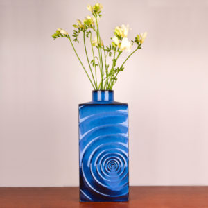 Das Design dieser schönen Vase stammt von dem Designer Cari Zalloni der in den 1960er Jahren mit seiner Arbeit als Designer begann. Er arbeitete für das Keramikwerk Steuler bei Coblenz. Die Vase ist vintage. Also original aus der Zeit. Ihr Zustand hervorragend. Die Farbe blau ist sehr edel. Das Design „Zyklon“ ist ein stilisierter Wirbelsturm, der sich in Wellen von der Mitte aus nach aussen bewegt und somit eine interessante Struktur auf der Vase bildet. Licht und Schatten wechseln sich ab. Die blaue Lasur bewegt sich zwischen hellerem und dunklerem Blau. Das leuchtende Königsblau strahlt jedoch auf der gesamten Vase. Die Vase hat eine rechteckige Form, wobei sie oben in einem engen Hals endet. Die Vase kannst Du als Tischvase und auch als Bodenvase verwenden. Durch ihre kraftvolle Ausstrahlung kann sie Beides sein. Ein echter Eye-Catcher. Du setzt damit ein Statement in Deinem zu Hause. Sei mutig und setze Akzente.