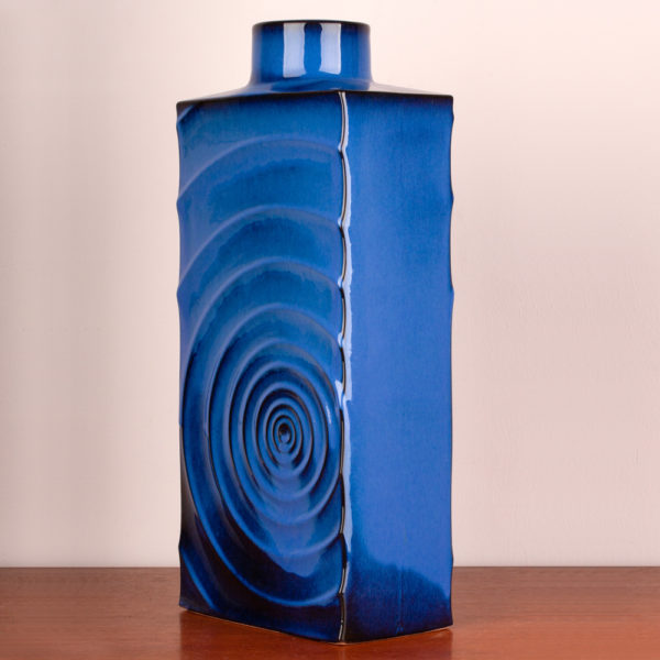 Das Design dieser schönen Vase stammt von dem Designer Cari Zalloni der in den 1960er Jahren mit seiner Arbeit als Designer begann. Er arbeitete für das Keramikwerk Steuler bei Coblenz. Die Vase ist vintage. Also original aus der Zeit. Ihr Zustand hervorragend. Die Farbe blau ist sehr edel. Das Design „Zyklon“ ist ein stilisierter Wirbelsturm, der sich in Wellen von der Mitte aus nach aussen bewegt und somit eine interessante Struktur auf der Vase bildet. Licht und Schatten wechseln sich ab. Die blaue Lasur bewegt sich zwischen hellerem und dunklerem Blau. Das leuchtende Königsblau strahlt jedoch auf der gesamten Vase. Die Vase hat eine rechteckige Form, wobei sie oben in einem engen Hals endet. Die Vase kannst Du als Tischvase und auch als Bodenvase verwenden. Durch ihre kraftvolle Ausstrahlung kann sie Beides sein. Ein echter Eye-Catcher. Du setzt damit ein Statement in Deinem zu Hause. Sei mutig und setze Akzente.