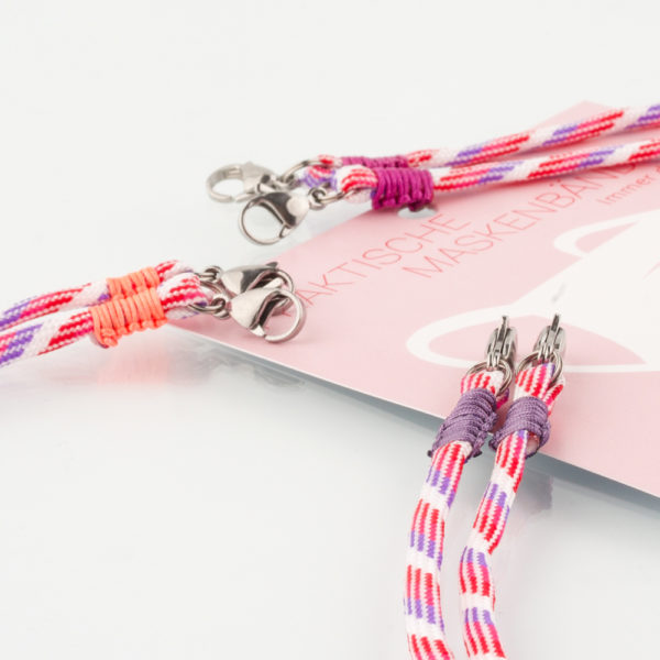 Das Maskenband ist der kleine Helfer im Alltag. Hier in der Farbkombi lila pink weiss und lila. Damit Du nicht ständig Deine Maske aus der Tasche holen musst, oder in der Hand hältst, hängst Du die Maske mit dem Band einfach um den Hals, oder steckst sie Dir in die Tasche Deines Oberhemdes, oder in die Tasche Deiner Jacke, wenn Du sie nicht benötigst. Sie kann aber auch einfach runterhängen. Ob Du bei der Arbeit bist, in ein Geschäft, oder ins Restaurant gehst, Du kannst sie schnell aufsetzen. Sogar mit einer Hand ist das möglich. Die zwei Karabinerverschlüsse klemmst Du einfach an das Gummiband, das bei Gebrauch um die Ohren gelegt wird. Durch das Eigengewicht hängen die Karabiner an der tiefsten Stelle des Gummibandes, sodaß sie auch nicht an den Ohren drücken können. Die Maskenbänder werden mit Deinem Farbwunsch angefertigt. Wähle Deine Wunschfarbe für die Bänder, die die Karabiner halten, im Menü aus.