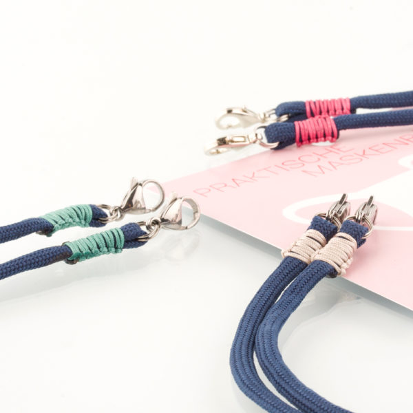 Das Masken Band ist der kleine Helfer im Alltag. Hier in der Farbkombi marineblau und beige. Eine klassische Farbe, die sich mit vielen Farben kombinieren lässt. Damit Du nicht ständig Deine Maske aus der Tasche holen musst, oder in der Hand hältst, hängst Du die Maske mit dem Band einfach um den Hals und steckst sie Dir in die Tasche Deines Oberhemdes, oder in die Revertasche Deiner Jacke, wenn Du sie nicht benötigst. Sie kann aber auch einfach runterhängen. Ob Du bei der Arbeit bist, in ein Geschäft, oder ins Restaurant gehst, Du kannst sie schnell aufsetzen. Sogar mit einer Hand ist das möglich. Die zwei Karabinerverschlüsse klemmst Du einfach an das Gummiband, das bei Gebrauch um die Ohren gelegt wird. Durch das Eigengewicht hängen die Karabiner an der tiefsten Stelle des Gummibandes, sodaß sie auch nicht an den Ohren drücken können. Die Maskenbänder werden mit Deinem Farb-Wunsch angefertigt. Wähle Deine Wunschfarbe für die Bänder, die die Karabiner halten, im Menü aus.