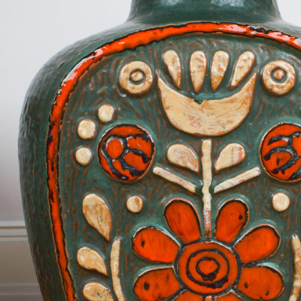 Die Bodenvase von der Firma Eduard Bay GmbH stammt aus den 1960er Jahren. Sie ist vintage. Also original aus der Zeit. Ihr Zustand ist sehr gut. Die Farben sind bunt zusammengesetzt. Olivgrün, rotorange, crémegelb und schwarz. Die florale ornamentartige Blumenkomposition erinnern an die Inka Zeit. Der Look ist archaisch. Wenn man so will, ist die Lasur der einzelnen Farben bewusst nicht flächendeckend ausgefüllt worden, sodaß die Farben auf der Oberfläche grob aneinander grenzen. Die Vase kannst Du als Bodenvase, aber auch als Tischvase verwenden. Gerade, bei der Größe der Vase, fällt sie optisch auf und ist ein Eye-Catcher und setzt damit ein Statement in Deinem zu Hause. Sei mutig und setze Akzente. Die Vase hat eine bauchige Flaschenform. Von unten schmal, basierend auf einer rechteckigen Grundform mit bauchigen Seiten, wird die Vase nach oben breiter und läuft wieder zusammen zu einem krempenartigen Hals, der in rotorange glasiert ist. Die Vase hat einen Kunsstoffeinsatz. Dieser wird bis heute für diese großen Vasen angeboten, damit man nicht so viel Wasser in die Vase füllen muss.