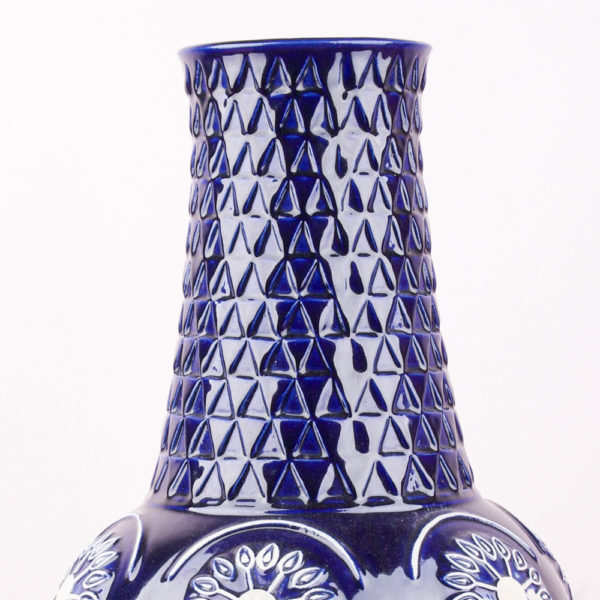 Die marineblaue und weisse Bodenvase steht auf einem Sockel der nach oben verlaufend in eine bauchige, fast kugelförmige Ausformung übergeht um darüber im gleichen Design in einen langen Vasenhals zur Öffnung übergeht. Die bauchige Ausformung besteht aus Romben mit einsetzten Kreisen und stilisierten Blumenblüten in weiss. Das Muster des Sockels und Halses ist in einer übereinanderliegenden Schuppentechnik in Dreiecksform gestaltet. Das Design stellt sich als Relief auf der Vase dar. Die Vase ist dunkelblau, glänzend lasiert und das Weiss der Keramik ist am Relief wie eine zweite Farbe zu sehen.