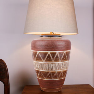 Die Tischlampe Boho Style Braun Creme hat eine warme Ausstrahlung. Die Farbe beruhigt und gibt dem Raum eine angenehme Atmosphäre. Der Lampenfuss besteht aus einer Vase aus den 1950er Jahren. Mittels einer Ritztechnik wurde in die Keramik ein geometrisches Muster in Ringen und senkrechten Linien aufgebracht und anschließend wurde die Vase teilweise mit weisser Farbe lasiert und gebrannt. Der oberer Rand der Vase ist mit einem kleinen Kragen in dunkelbraun gestaltet. Der Schirm und die Elektrik sind oben in den Hals der Vase eingesteckt und lassen sich bei Gebrauch der Vase abnehmen und auch als Blumenvase nutzen. Diese Lampe ist ein Unikat.