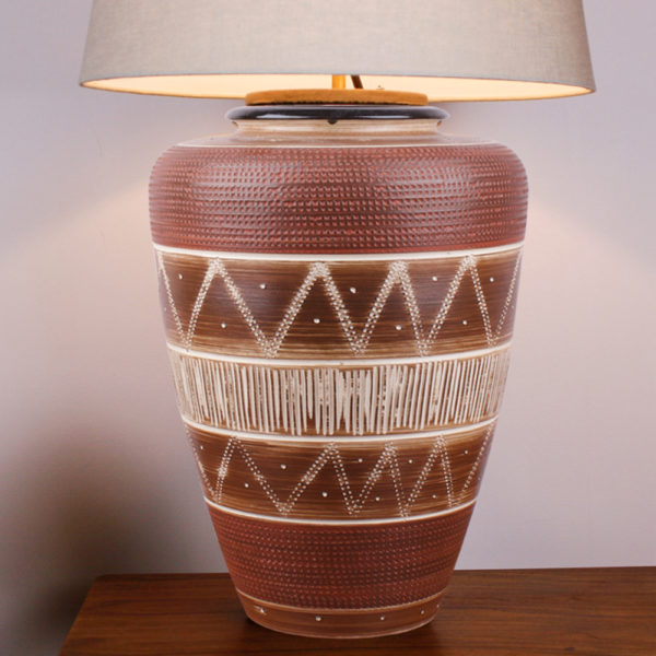 Die Tischlampe Boho Style Braun Creme hat eine warme Ausstrahlung. Die Farbe beruhigt und gibt dem Raum eine angenehme Atmosphäre. Der Lampenfuss besteht aus einer Vase aus den 1950er Jahren. Mittels einer Ritztechnik wurde in die Keramik ein geometrisches Muster in Ringen und senkrechten Linien aufgebracht und anschließend wurde die Vase teilweise mit weisser Farbe lasiert und gebrannt. Der oberer Rand der Vase ist mit einem kleinen Kragen in dunkelbraun gestaltet. Der Schirm und die Elektrik sind oben in den Hals der Vase eingesteckt und lassen sich bei Gebrauch der Vase abnehmen und auch als Blumenvase nutzen. Diese Lampe ist ein Unikat.