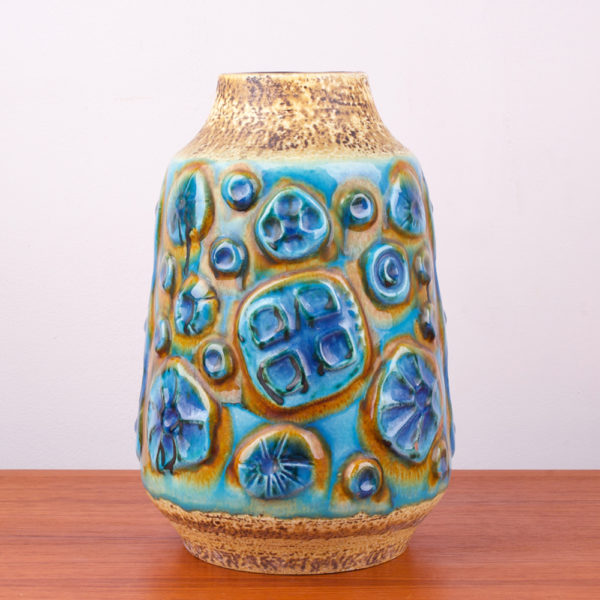 Diese Vase hat so erfrischende Farbkomponenten, dass man gleich an Meer, Sand und Himmel denken muss. Sie läuft konisch, von unten breit zum Hals schmal zu. Der Hals der Vase setzt sich vom Korpus durch einen Schwung nach oben ab. Das wilde Dekor auf der Vase erinnert an ein archaisches Muster, oder auch an stilisierte Blütenstände. Oder auch an Versteinerungen. Die sandfarbene Oberfläche der Vase ist unregelmäßig, rauh und die blauen und türkis lasierten Flächen sind hochglänzend gestaltet. Die Oberfläche wechselt sich somit sehr interessant ab. In der Mid Century Ära wurde in der Gestaltung das archaische Thema gern interpretiert. Der türkisfarbene Ton erinnert an karibisches Meer. Die sandfarbene Oberfläche ist wie Sand am Meer. Diese Vase ist ein echtes Statement und braucht zum Ausgleich zum Beispiel einen Teakholztisch. In die Vase passen ganz wunderbar Pampasgras, oder hochstilige Blumen.