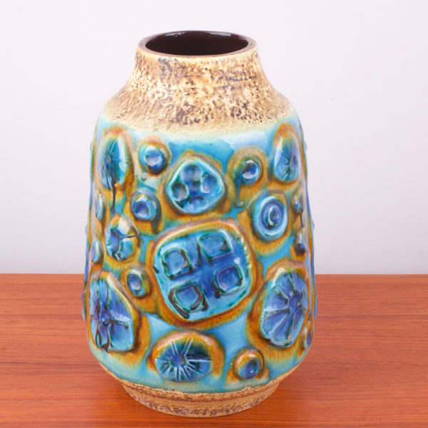 Diese Vase hat so erfrischende Farbkomponenten, dass man gleich an Meer, Sand und Himmel denken muss. Sie läuft konisch, von unten breit zum Hals schmal zu. Der Hals der Vase setzt sich vom Korpus durch einen Schwung nach oben ab. Das wilde Dekor auf der Vase erinnert an ein archaisches Muster, oder auch an stilisierte Blütenstände. Oder auch an Versteinerungen. Die sandfarbene Oberfläche der Vase ist unregelmäßig, rauh und die blauen und türkis lasierten Flächen sind hochglänzend gestaltet. Die Oberfläche wechselt sich somit sehr interessant ab. In der Mid Century Ära wurde in der Gestaltung das archaische Thema gern interpretiert. Der türkisfarbene Ton erinnert an karibisches Meer. Die sandfarbene Oberfläche ist wie Sand am Meer. Diese Vase ist ein echtes Statement und braucht zum Ausgleich zum Beispiel einen Teakholztisch. In die Vase passen ganz wunderbar Pampasgras, oder hochstilige Blumen.